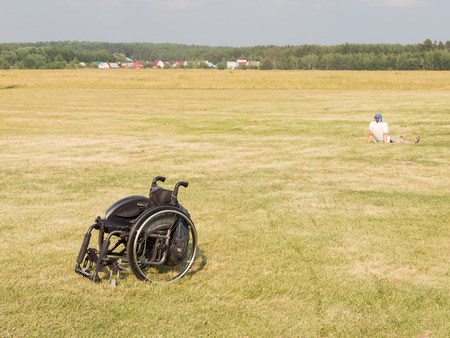 The Moscow region - 15 July 2016: Black wheelchair standing in a field on festevale "Sky for all" and the man away sitting on green grass mown July 15, 2016, Moscow Region, Russiaのeditorial素材