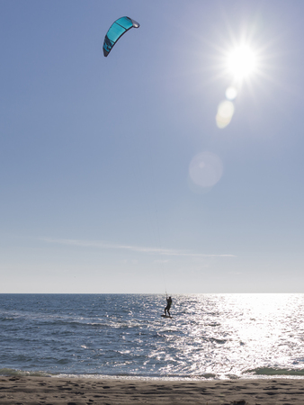 The unrecognizable kitesurfer rides on sea water on a board with a blue kite in bright sunlight and glare on the waterの写真素材