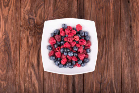 Raspberries with blueberries in a white bowl on a brown wooden background top viewの写真素材