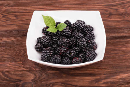 White bowl filled with blackberries with leaves on a brown wooden backgroundの写真素材