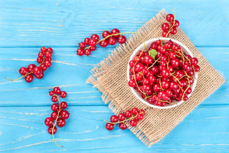 Bowl full of fresh red currant  with scattered berries on the sacking and wooden background, top...