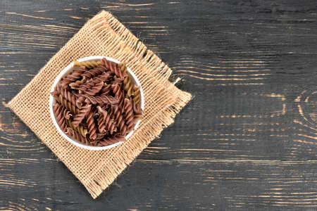 Uncooked pasta fusilli in a bowl on burlap and table, top viewの写真素材