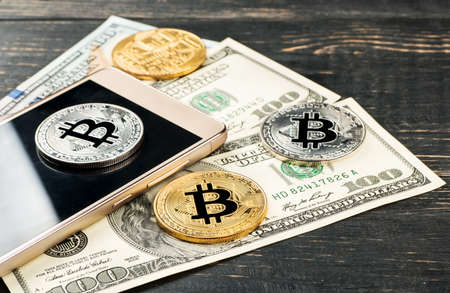 Gold and silver bitcoin coins with dollars and smartphone on wooden backgroundの写真素材