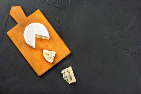 Brie cheese with two slices on cutting board on dark wooden background, top view