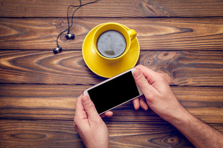 Mockup image of a mobile phone with an empty black screen and coffee in a yellow cup on an old wooden table in a cafe.の写真素材
