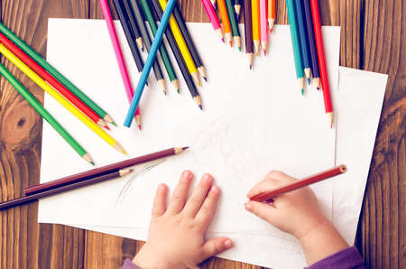 The child's hands are painted with colored pencils on a white sheet of paper on a wooden table.