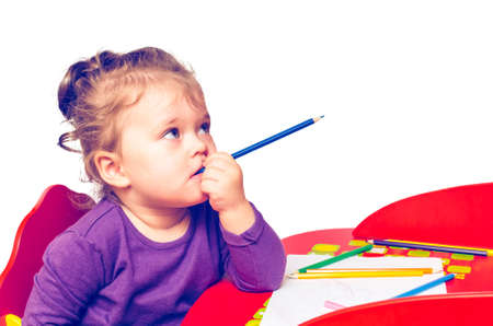 Little girl dreams and bites a pencil while sitting at a table, isolated on a white background.の写真素材