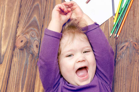 Laughing little girl lies on a wooden floor with scattered colored pencils.の写真素材