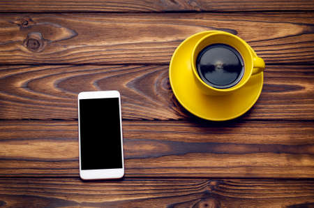 Yellow cup with coffee and phone on a wooden table.の写真素材