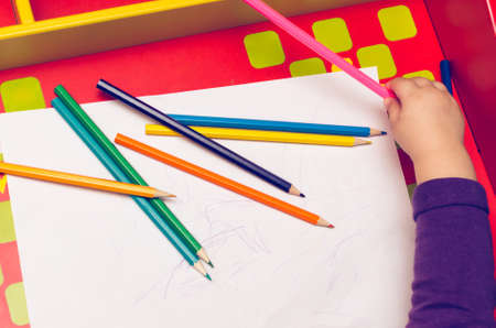 On the red table, children's hands with colored pencils and a blank sheet of paper.の写真素材