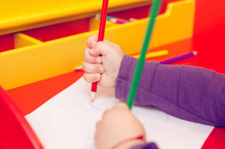 On the red table, children's hands with colored pencils and a blank sheet of paper.