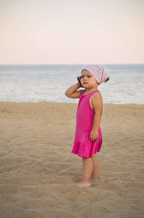 A little girl in a red dress is talking on the phone on the sea beach.の写真素材