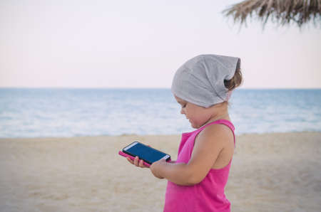 little girl in a red dress holding a phone in her hands on the sea beach.の写真素材