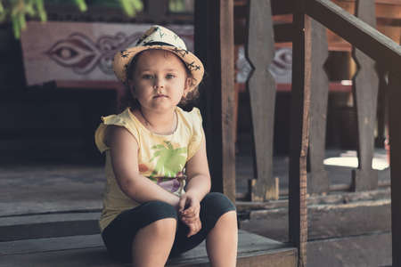 little girl in a hat sits on the porch of an old wooden house.の写真素材