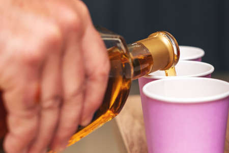 A man's hand holds a glass bottle with alcohol and pours an alcoholic drink into paper cups.の写真素材