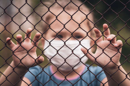 A child in a medical mask holds the fence with his hands. Quarantine melancholy and loneliness concepts.の写真素材