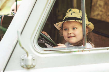 Little girl in a hat sits behind the wheel of an old disassembled car.