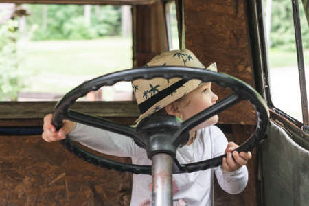 Little girl in a hat sits behind the wheel of an old disassembled car.