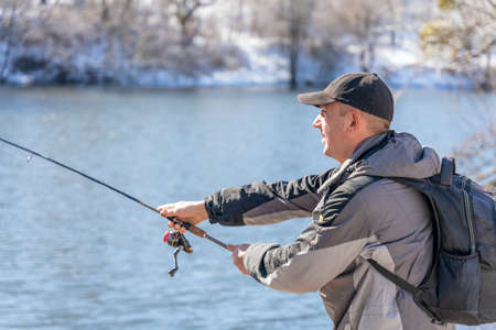 A fisherman with a fishing rod and a backpack catches fish on the bank of a snow-covered river in early spring.の写真素材