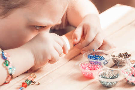 A little girl is engaged in needlework, making jewelry with her own hands, stringing multi-colored beads on a thread.の写真素材