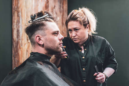 Girl hairdresser makes a haircut to a young guy using a hair clipper in a barbershop.の写真素材