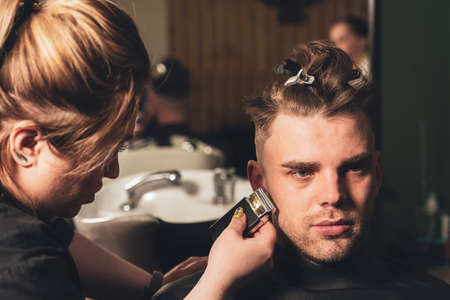 Girl hairdresser makes a haircut to a young guy using a hair clipper in a barbershop.