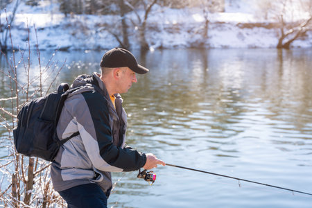 A fisherman with a fishing rod and a backpack catches fish on the bank of a snow-covered river in early spring.の写真素材
