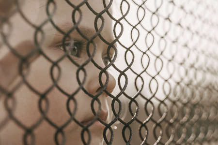 A little girl with a sad look behind a metal fence. Social problem of refugees and forced migrants.の写真素材