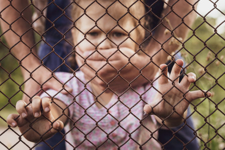 The child's hands are holding onto a metal mesh fence. The problem of forced deportation of Ukrainian children from the territories occupied by Russia.の写真素材