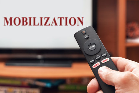 A man's hand holds a TV remote control, all buttons on the remote control are signed news, on the TV there is an inscription mobilization.の写真素材