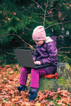 A cute little girl sits on an old stump in the middle of the forest and uses a laptop for online learning or playing games.の写真素材