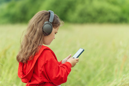 Cute little girl in wireless headphones holding smartphone and enjoying music outdoors.の写真素材