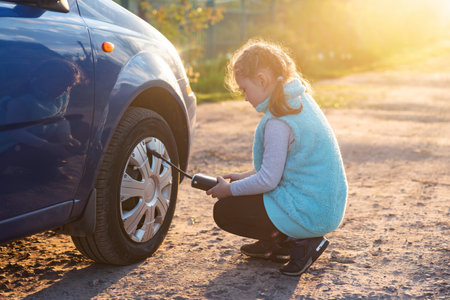 A little girl pumps up a car wheel with a compressor at sunset on a dirt road.の写真素材