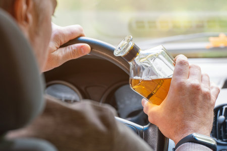 The driver is sitting behind the wheel and holding a bottle of alcoholic drink. Breaking the law and drinking alcohol while driving.の写真素材