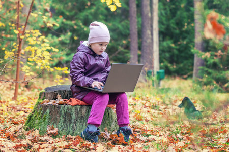 A cute little girl sits on an old stump in the middle of the forest and uses a laptop for online...