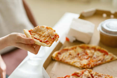 A woman's hand takes a piece of hot fresh pizza cut into slices in a cardboard package on the table.の写真素材