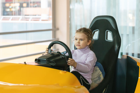 A little girl in an entertainment center with slot machines sits in a car game simulator and holds the steering wheel with her hands.の写真素材