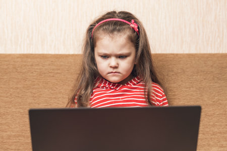 Beautiful little girl with serious expression using laptop for entertainment or education sitting on...