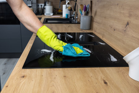 Close up of hand in yellow glove cleaning modern induction hob in kitchen