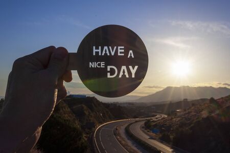 A hand holds a metal disk with the inscription âHave a nice dayâ cut out, on a blurred background of a highway surrounded by mountains during sunriseの写真素材