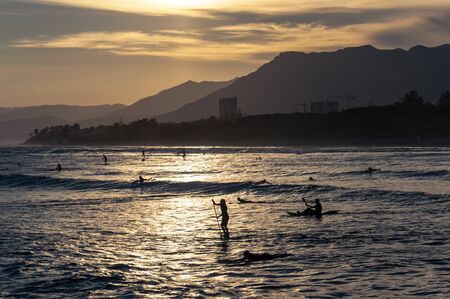 Girl on a board with a paddle and a guy on a kayak on the sea with waves in the light of the setting sun on the background of the coastline with palm trees, a building and mountains.の写真素材