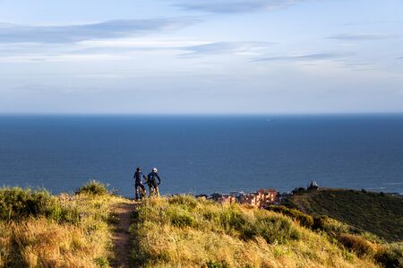 Two cyclists on a mountain road from the top of the hill look at the sea and the village below on...