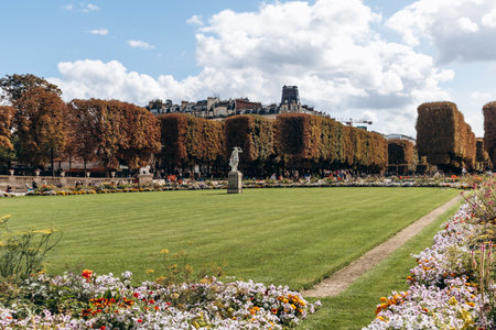 A scenic view of the Luxembourg Gardens with manicured lawns, blooming flowerbeds, and classical statues surrounded by tall autumn-colored trees, offering a peaceful atmosphere in the heart of Paris.の写真素材