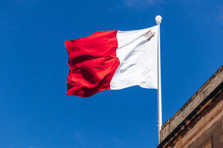 The national flag of Malta, red and white with the George Cross in the upper hoist corner, flies against a clear blue sky above traditional Maltese limestone architectureの写真素材