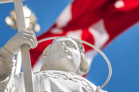 White statue of a saint holding a cross stands against a vivid blue sky, dramatically framed by the bold red-and-white flag of the Knights of St John waving overhead in a traditional Maltese settingの写真素材