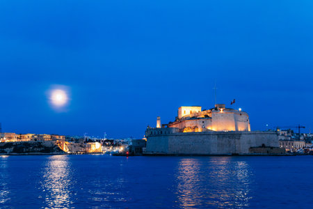 Illuminated Fort St. Angelo stands on the Grand Harbour at twilight in Malta, its limestone bastions glowing beneath a blue evening sky and full moon, with reflections shimmering on the water.の写真素材
