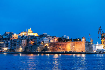 Panoramic twilight view of Vallettaâs historic waterfront with illuminated bastions, limestone buildings, and a church dome glowing above the Mediterranean Seaの写真素材