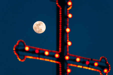 A glowing full moon rises behind a large decorative cross adorned with red lights, part of the festive street decorations for a traditional Maltese village celebrationの写真素材