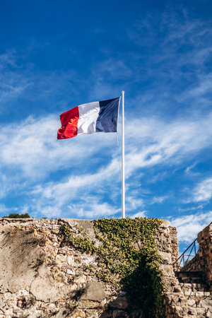 Roquebrune Cap Martin, France - February 9, 2025: French national flag flying above the stone walls of Grimaldi Castle against blue skyの写真素材