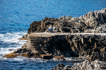 Roquebrune-Cap-Martin, France â 9 February 2025: A woman sits on a rocky seaside platform reading a book with her dog beside her, surrounded by rugged cliffs and deep blue Mediterranean waters on a sunny dayのeditorial素材